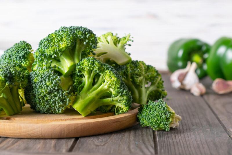 on a rustic looking wooden surface is a round wooden chopping board with broccoli florets on it, beside it are loose broccoli florets, at the back is a couple of green bell peppers and garlic cloves