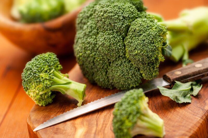 on a wooden surface is a wooden chopping board with broccoli head, florets, and knife on it,  at the back is a wooden bowl with broccoli florets