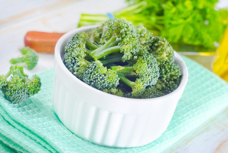 a closeup image of a white ceramic bowl full of broccoli florets resting on a light green table napkin