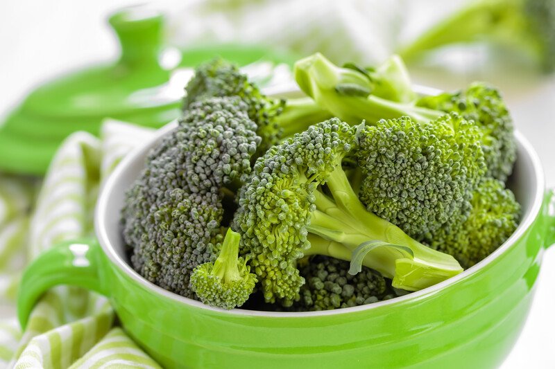 a closeup image of a ceramic green bowl with ears full of broccoli florets with a green and white kitchen towel beside it and green ceramic cover at the back