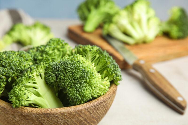 a closeup image of a wooden bowl full of broccoli florets, at the back is a wooden chopping board with broccoli florets and a knife