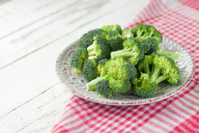 on a white wooden surface is a partially folded red and white table cloth with a plate on it with broccoli florets