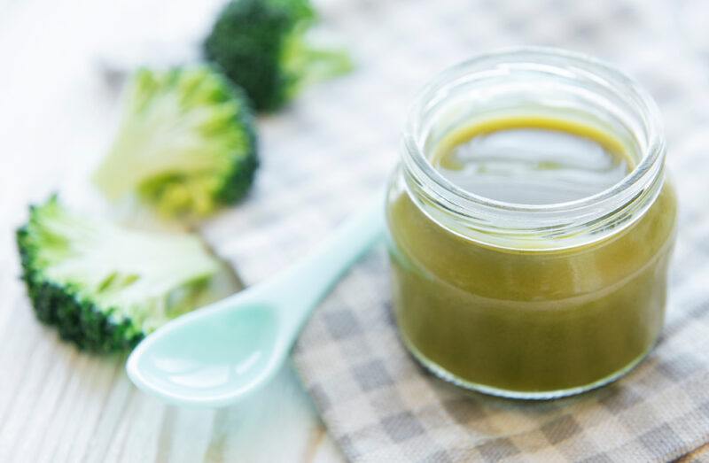 on a wooden surface is a small jar with broccoli puree resting on top of a striped white and blue table napkin, beside it is a plastic baby spoon and three broccoli florets