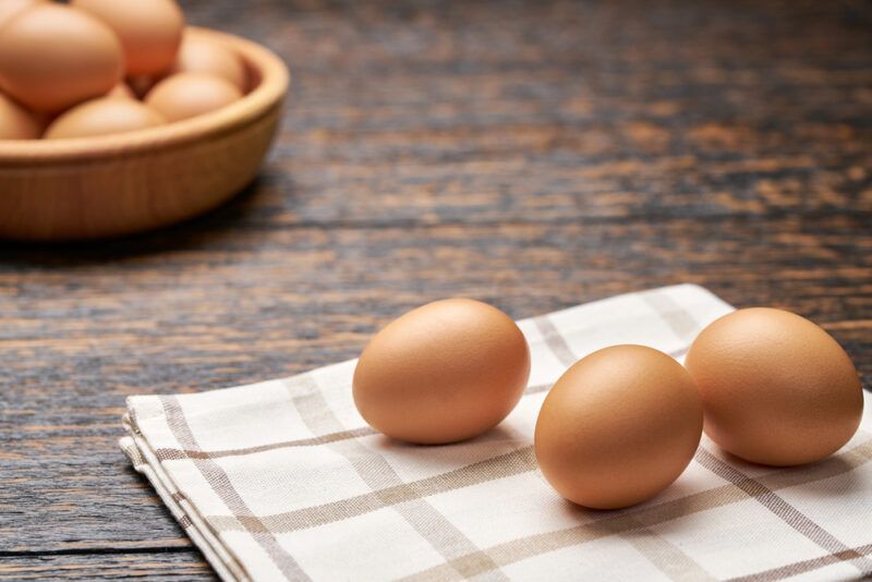 on a dark rustic-looking wooden surface are three brown eggs resting on top of a white table napkin with brown stripes, at the back is a wooden dish full of brown eggs