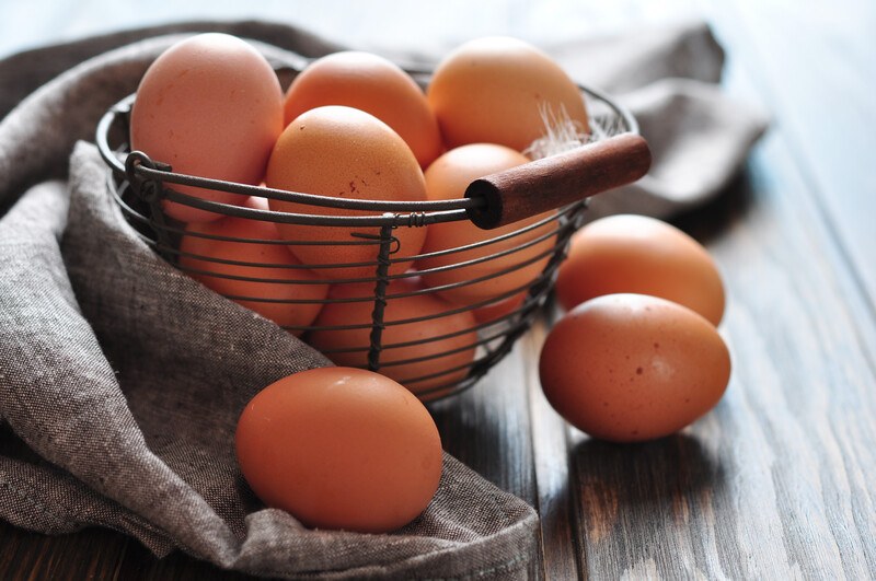 on a wooden surface is a wire basket full of brown eggs with a grey table napkin behind it and 3 loose brown eggs around it