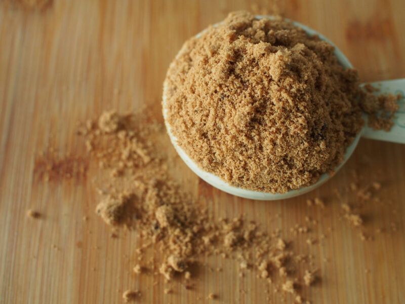 on a wooden surface is a white measuring spoon full of brown sugar with loose brown sugar around it