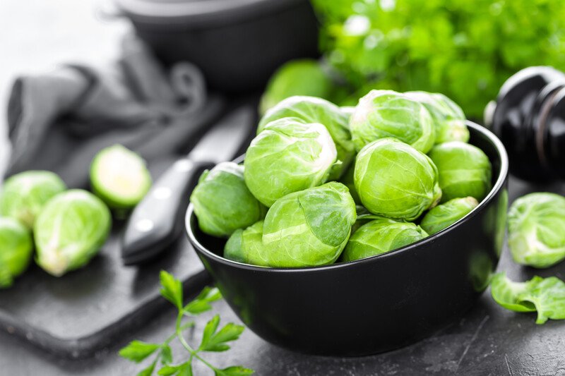 a black bowl full of brussels sprouts with brussels sprouts and greens at the back with knife and black chopping board