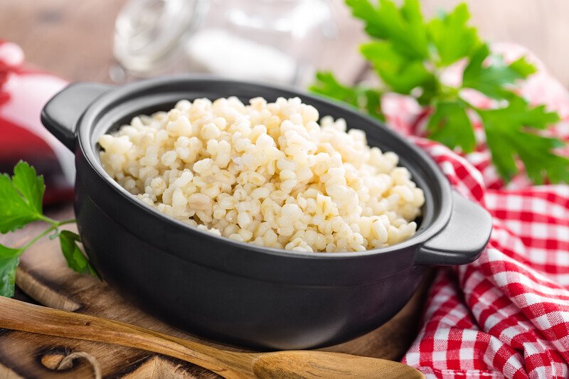 a black bowl full of cooked bulgur, with a red and white table napkin, fresh herbs, and wooden spoon beside it