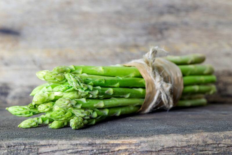 on an aged wooden surface is a bunch of fresh asparagus tied with a brown string over a narrow brown paper