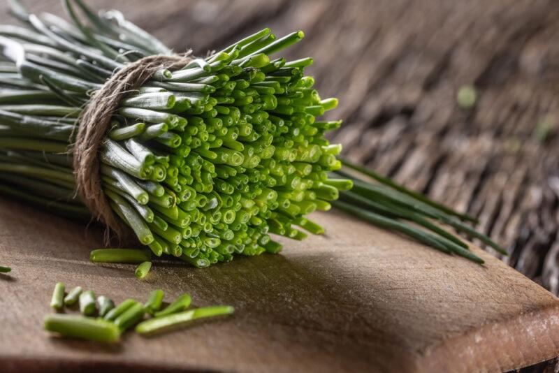 on a rustic looking wooden surface is a wooden chopping board with a bunch of chives partially minced