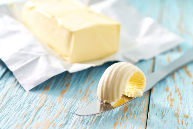 on a blue rustic wooden surface is a block of butter sitting on top of its wrapper, in front of it is a  butter knife with butter on it