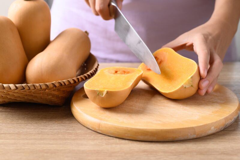 on a wooden surface is a partial image of a woman cutting butternut squash in half with a knife on a wooden chopping board, beside it is a basket of whole butternut squash