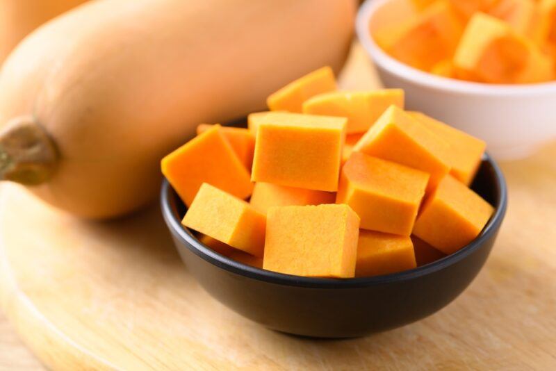 on a wooden surface is a dark brown ceramic bowl and white bowl full of cubed butternut squash, with a whole butternut squash at the back