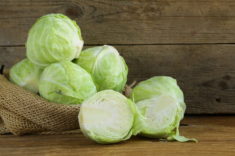 a burlap sack with green cabbages in it and a couple cabbages outside the burlap sack, one cut in half, resting on a wooden surface with wooden back wall as well