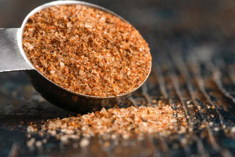 on a dark wooden surface is a metal measuring spoon slightly tilted, full of cajun spice, with some of its content on the surface 