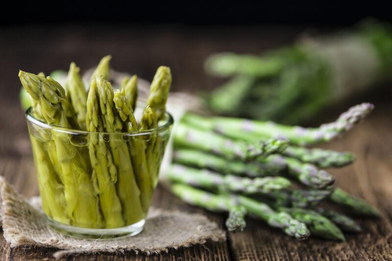 on a wooden surface is a preserved asparagus in a jar resting on top of a piece of burlap, at the back are a couple of bundles of fresh asparagus