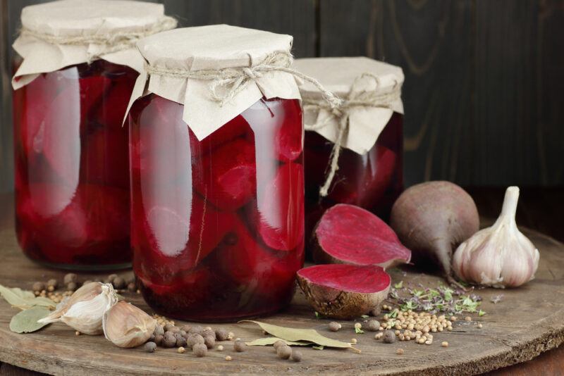 on a wooden surface are three jars of canned beets, around them are halve and whole fresh beets, garlic head, garlic cloves, laurel leaves, and loose spices and herbs