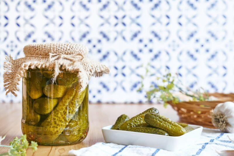 on a wooden surface with white and blue prints background is a jar of canned cucumber with a square dish of pickled cucumber resting on top of a white table napkin with blue stripes with a basket with sprigs of greens and a garlic head beside it