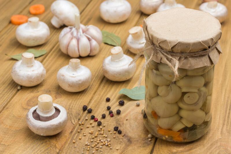 on a wooden surface is a jar of canned button mushrooms with loose fresh button mushrooms, spices, and head of garlic around it