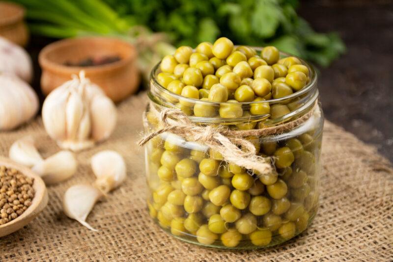 on a wooden surface is a jar full of canned peas resting on top of a burlap, with a head and cloves of garlic, spices, and herbs beside it