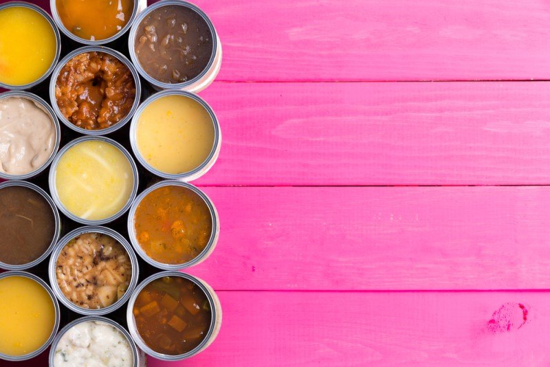 This photo shows an overhead view of several types of canned soups with the lids open, on a pink wooden surface.