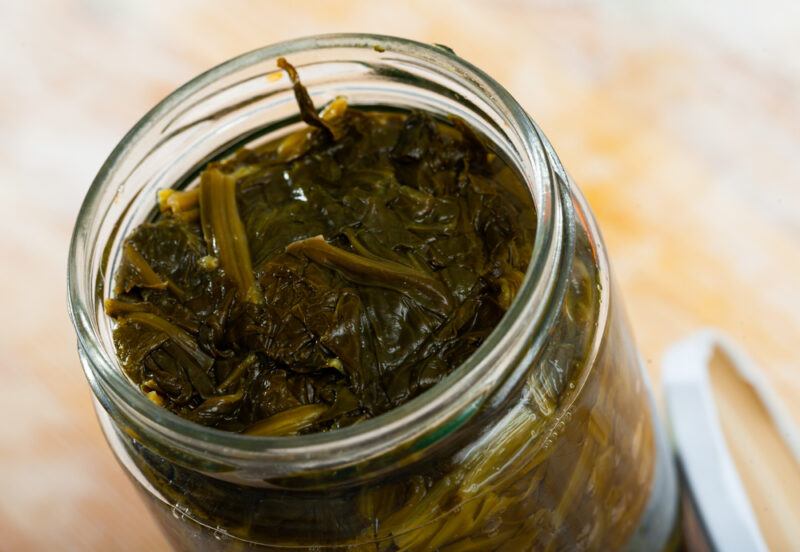 a closeup image of the mouth of a jar of canned spinach with its cover leaning beside the bottle