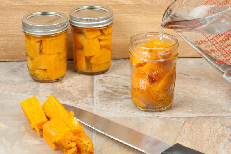 three mason jars with sweet potatoes with one being poured liquid into,  pieces of cubed sweet potatoes and a knife can be seen in the foreground