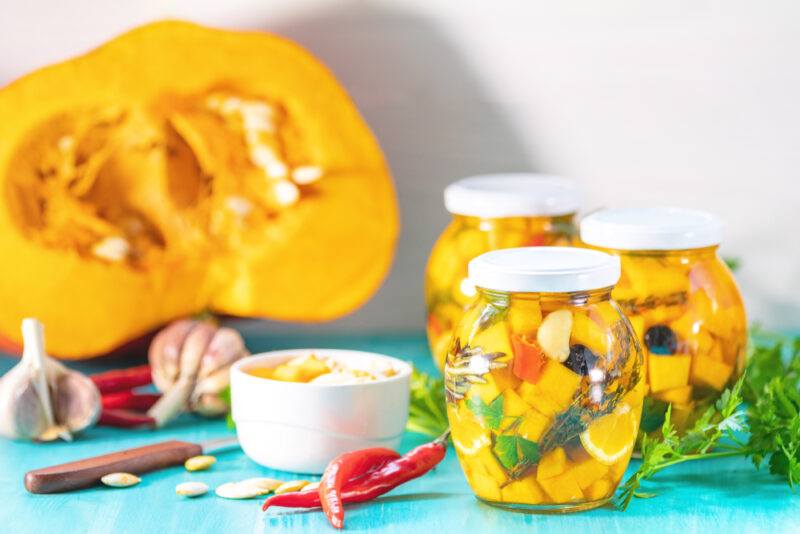 on a sky-blue surface are a spread of an open squash with seed showing, heads of garlic, a knife, fresh chili peppers, herbs, and three jars of canned winter squash