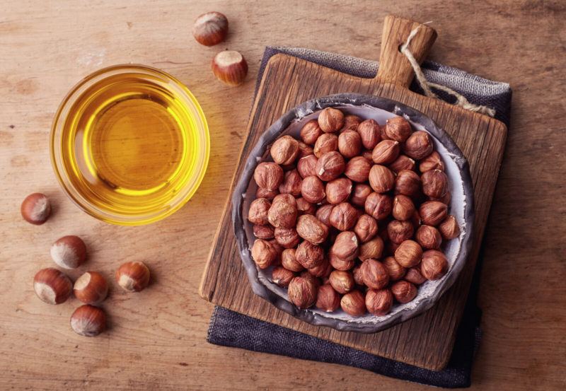 bowl of hazelnuts with a container of hazelnut oil beside it | Food For Net a glass cup with some hazelnut oil, placed beside a bowl filled with raw hazelnuts