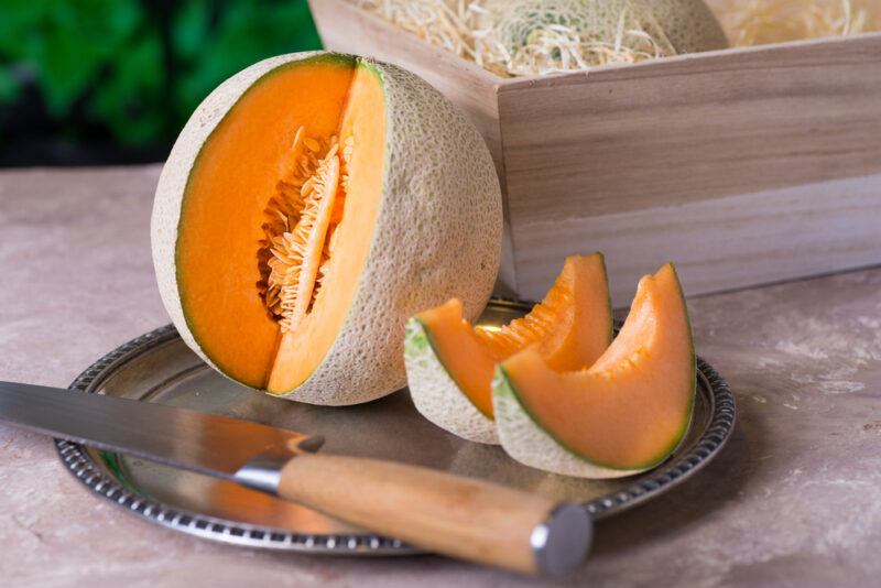 on a grey surface is a metal platter with partially cut cantaloupe with a knife beside it, at the back is a wooden crate with whole cantaloupes