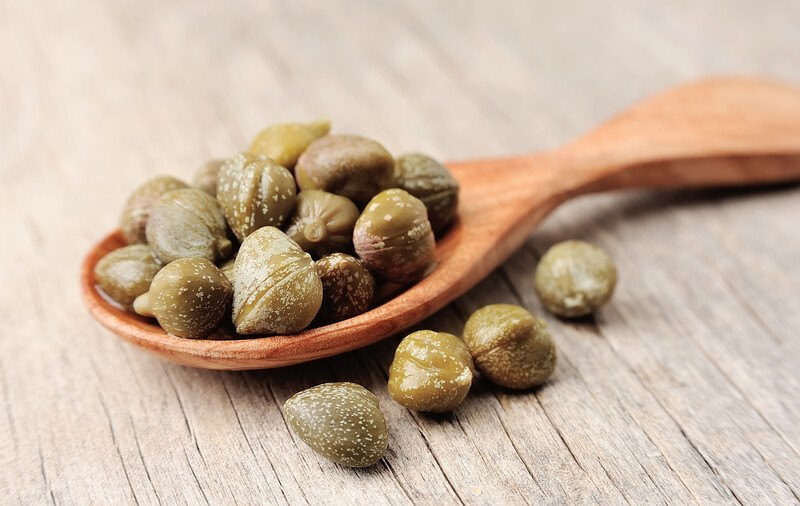 on a wooden surface is a closeup image of a wooden spoon full of capers, with loose capers beside it