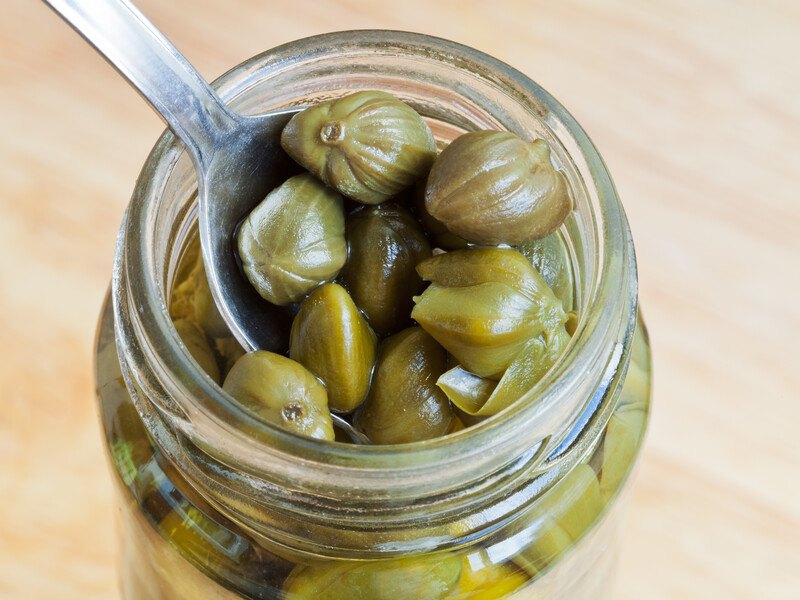 a closeup image of a wooden surface with a small jar of capers with a silver spoon in it