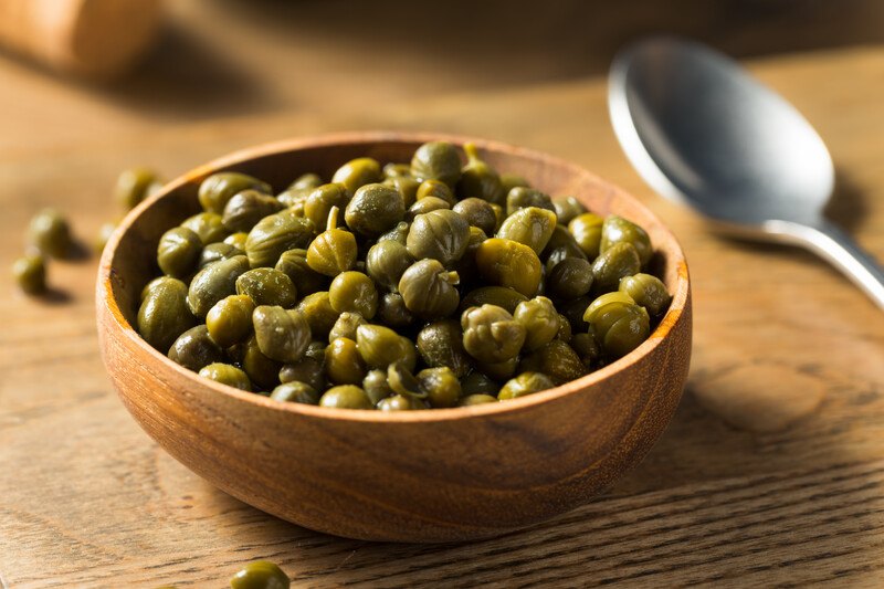 on a wooden surface is a wooden bowl full of capers, beside it is a silver spoon and loose capers around it