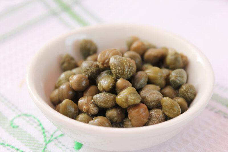 a closeup image of a white ceramic bowl with capers resting on a white table cloth with green stripes