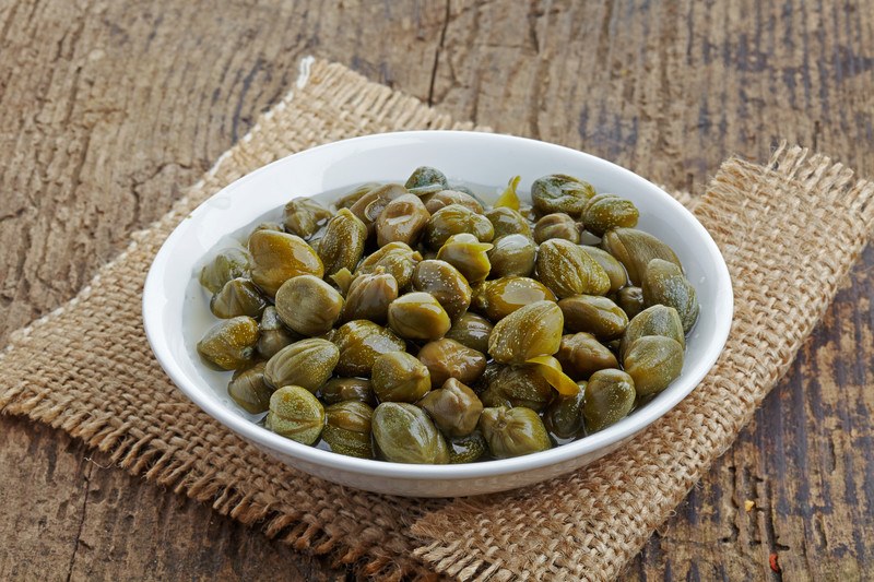 on a wooden surface is a white shallow bowl with capers resting on a burlap