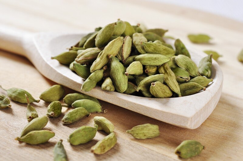 a closeup image of a wooden ladle full of cardamoms with loose cardamom around it