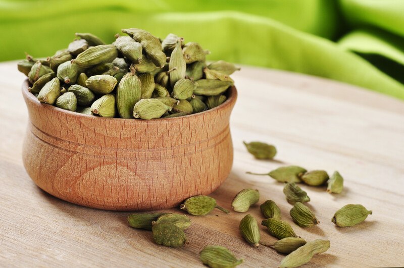 closeup image of a wooden bowl full of cardamom pods with loose cardamom around it