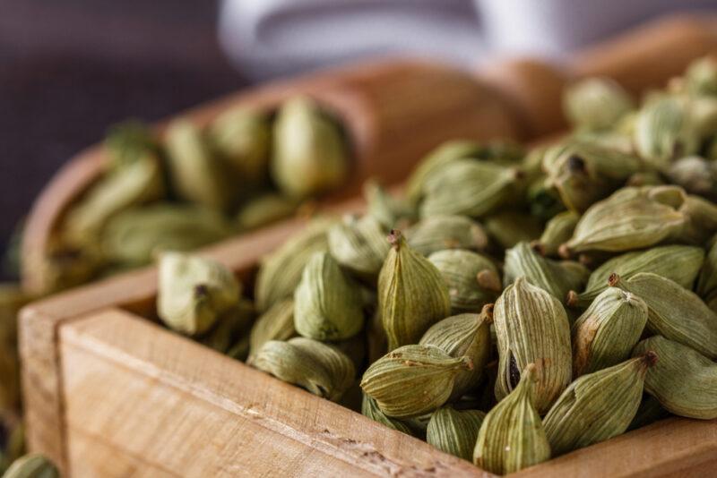 closeup image of cardamom in a square wooden crate with a wooden scoop full of cardamom pods