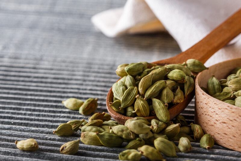 on a dark grey place mat is a wooden spoon resting beside a partially visible bowl, both filled with cardamom, around them are loose cardamom pods