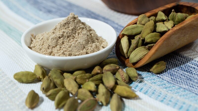 on a white and blue table cloth is a small white dish with ground cardamom with wooden scoop full of cardamom with some spilling out from it