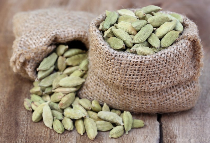 cardamom pods in a a couple of tiny brown sacks resting on a wooden surface; one standing upright, one on its side with cardamom pods pouring out