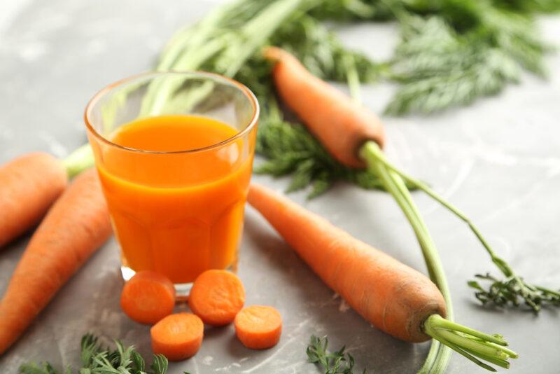 on a concrete surface are fresh carrots and a clear glass of carrot juice with sliced carrots beside it