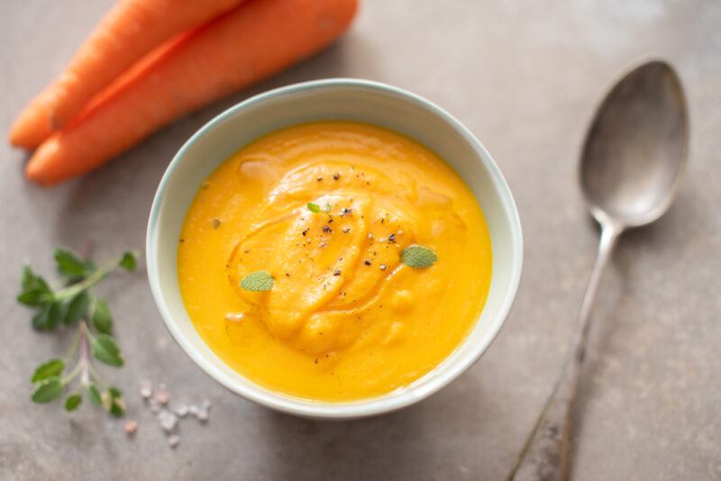 on a concrete surface is a white bowl full of carrot puree, beside it is a couple of carrots, sprigs of cilantro, and a silver spoon