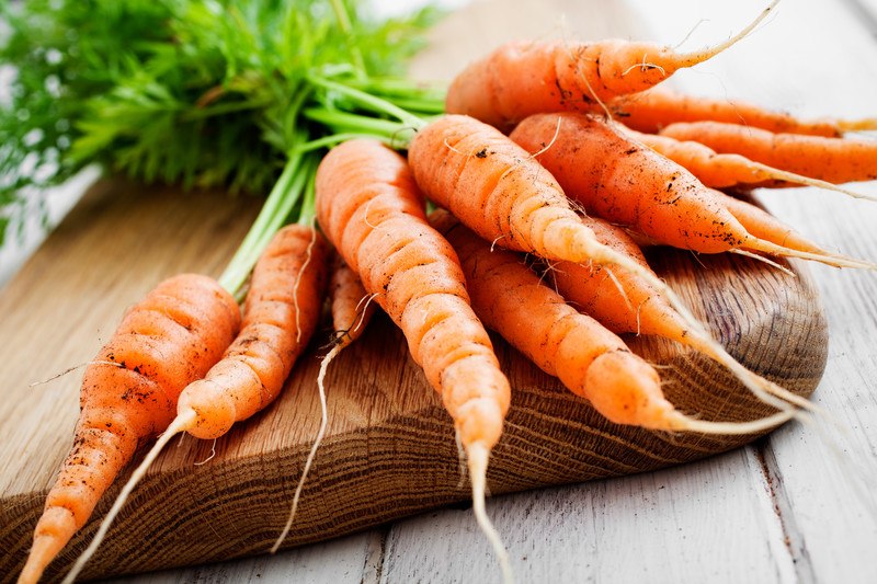 closeup image of a bunch of carrots with leaves still attached, laid out on a wooden chopping board, on a wooden surface