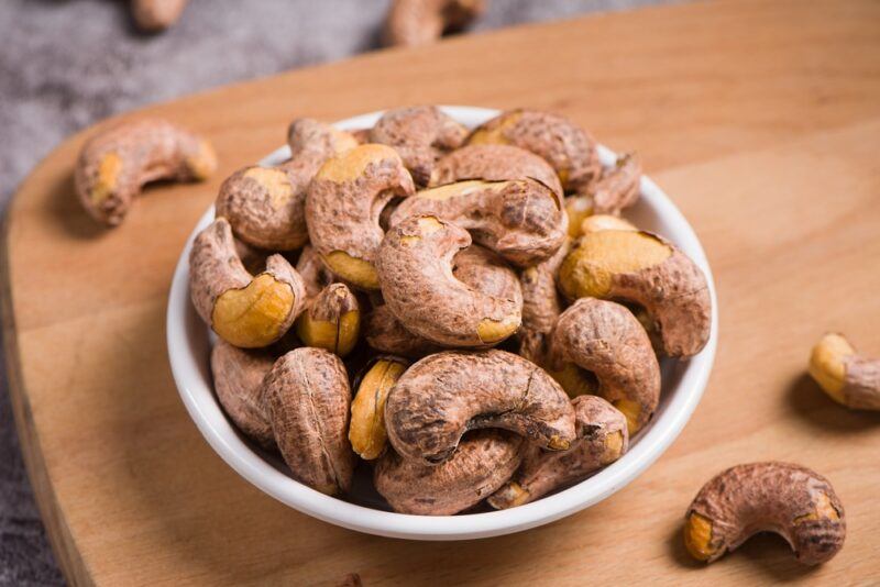 on a marbled surface is a wooden chopping board with a small bowl of cashew nuts, around it are loose cashew nuts