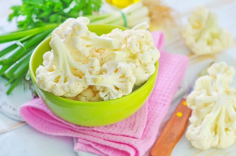 a mint green bowl full of cauliflower florets, resting on a pink table napkin with florets beside it and a knife, at the back is some greens as well