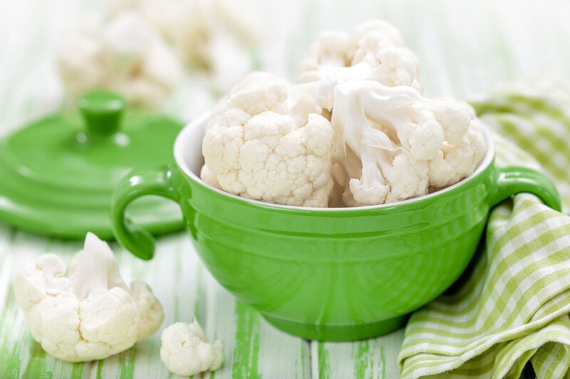 green ceramic bowl with ears full of cauliflower florets with a white and green kitchen towel with loose cauliflower florets around it and green ceramic cover