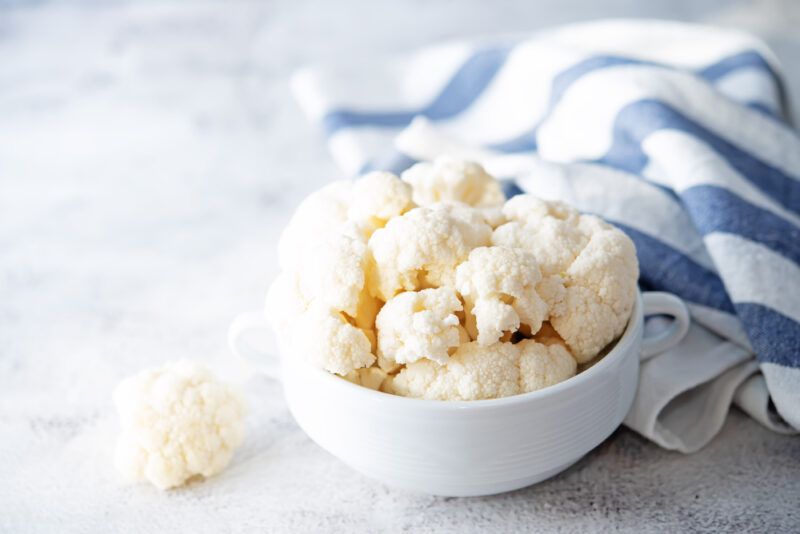 on a marble surface is a white dish full of cauliflower florets with a loose floret beside it, at the back is a white and blue kitchen towel