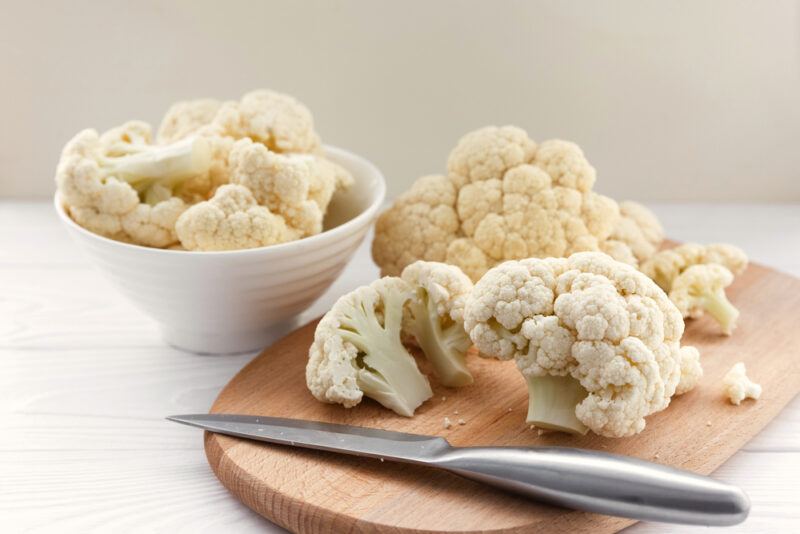 on a white wooden surface is a brown wooden chopping board with cauliflower florets and a knife, the white bowl behind it is also full of cauliflower florets