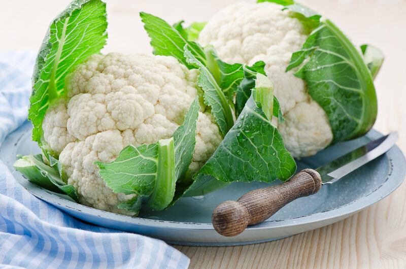 on a light wooden surface is a blue plate with a couple of cauliflower heads and a knife, beside it is a blue and white table napkin